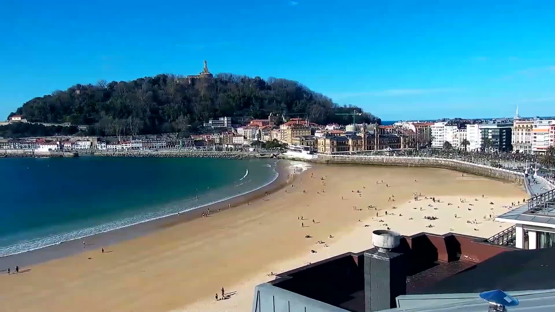 A sandy beach with scattered people is curved around a bay, with a town and a hill rising behind it under a clear blue sky.