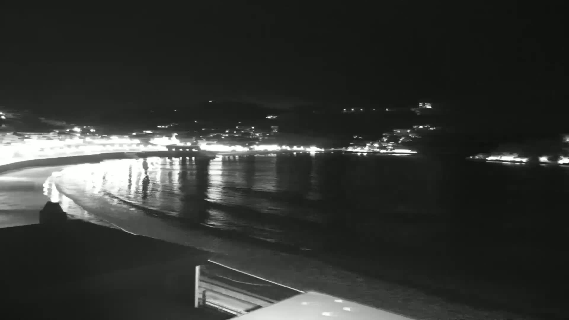 A nighttime view of a calm ocean reflecting the lights of a town nestled against hills.