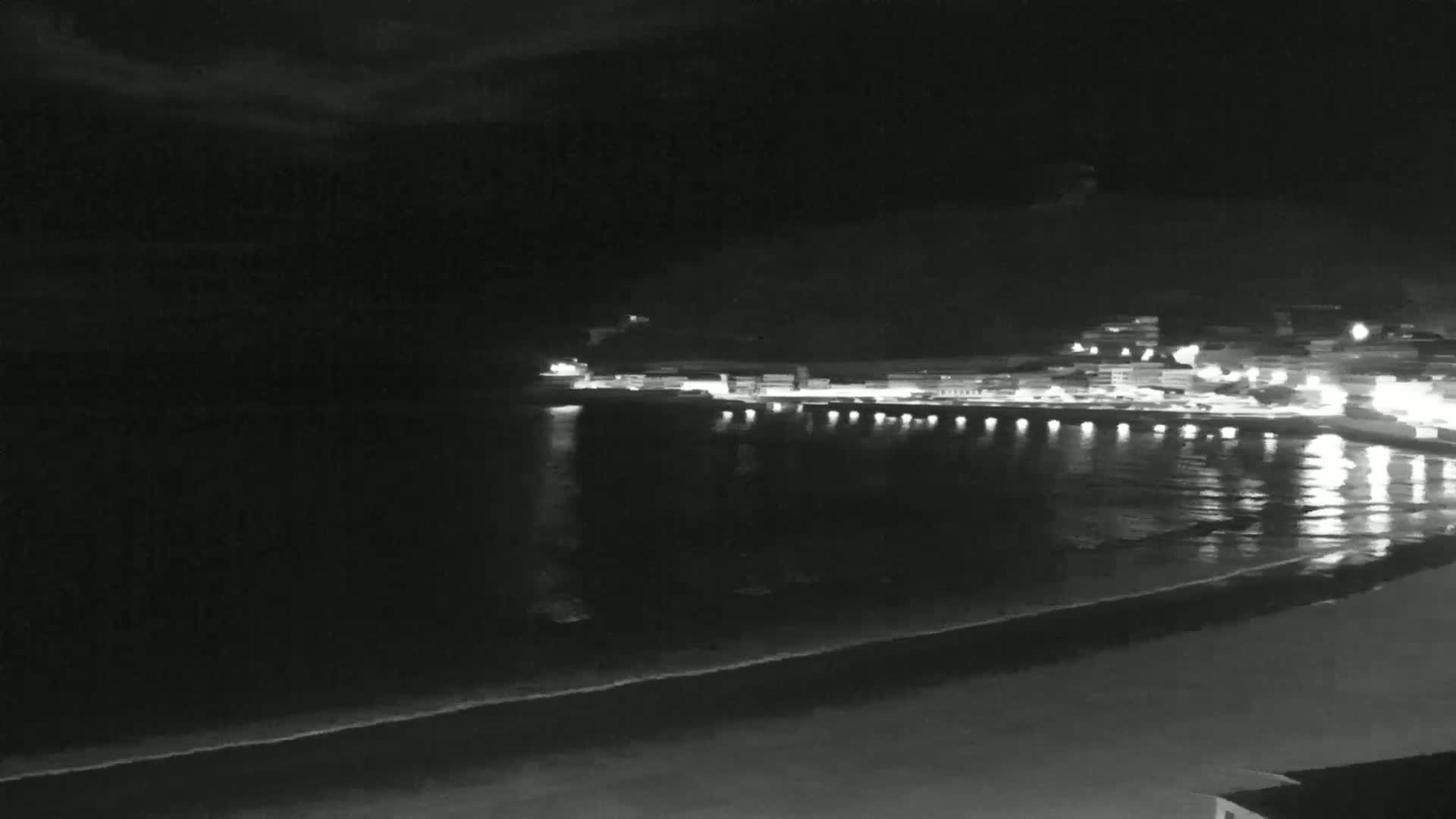 A nighttime coastal scene shows a dark beach, calm water reflecting the lights of a town on a hill in the background.