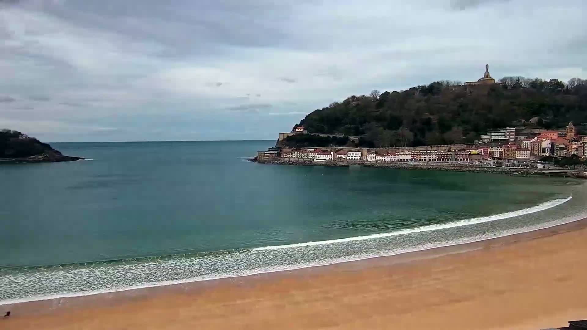A sandy beach curves around a calm bay under a cloudy sky, with a town nestled against a hill featuring a tall monument in the background.