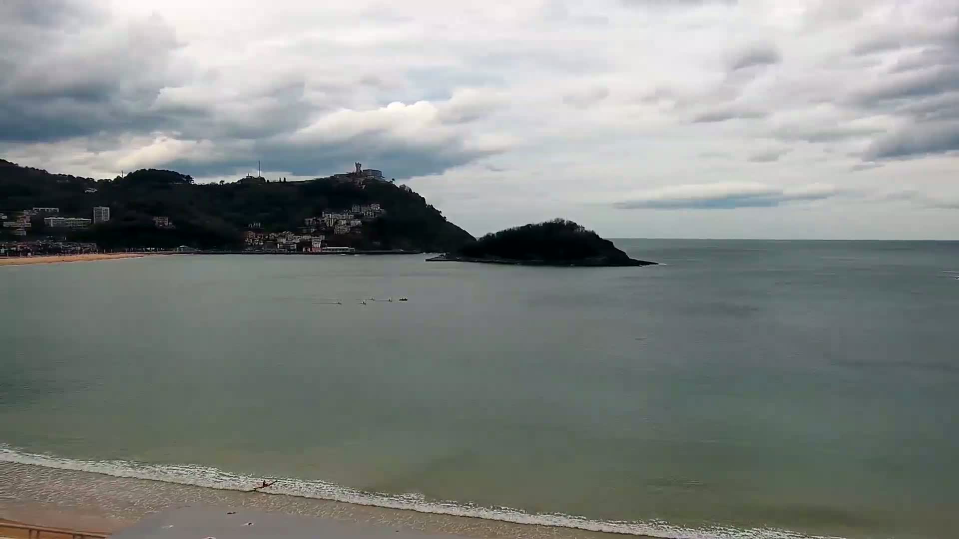 A calm bay with a sandy beach and a few people in the water is overlooked by a hill with buildings on a cloudy day.