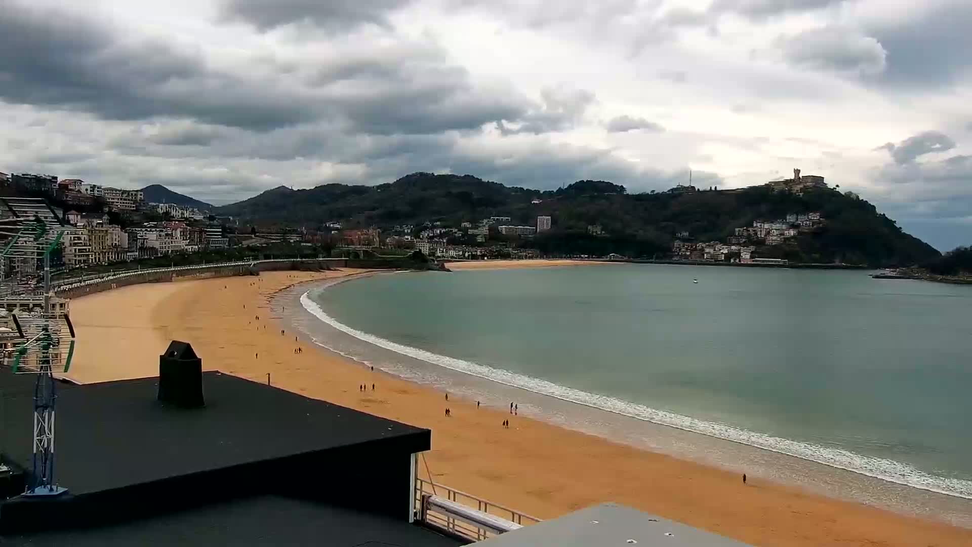 A sandy beach curves around a calm bay under a cloudy sky, with buildings and hills rising in the background.
