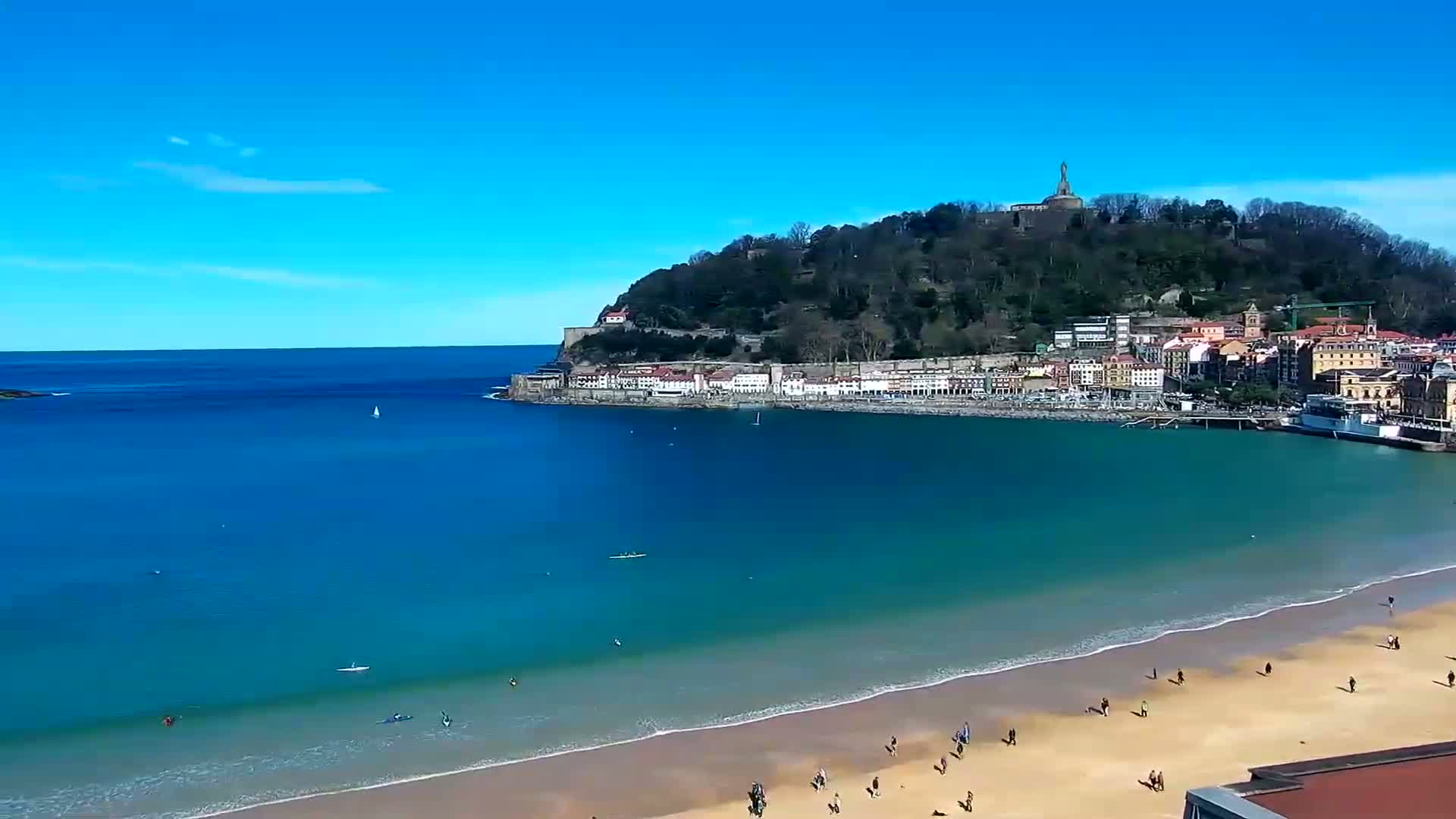 A sandy beach with people on it and calm, blue water laps the shore near a town nestled at the base of a green hill topped with a statue under a clear blue sky.