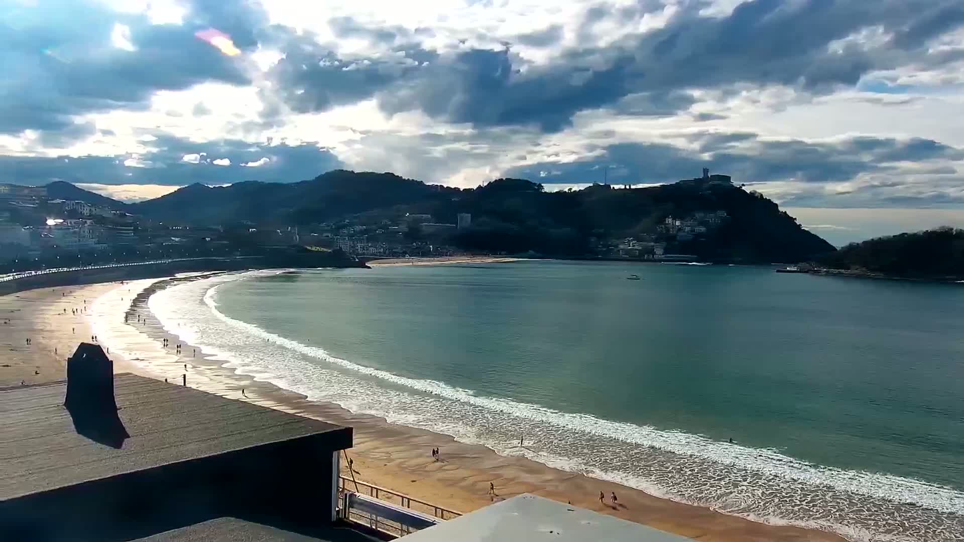 A long, sandy beach curves around a bay, with gently rolling waves under a partly cloudy sky.