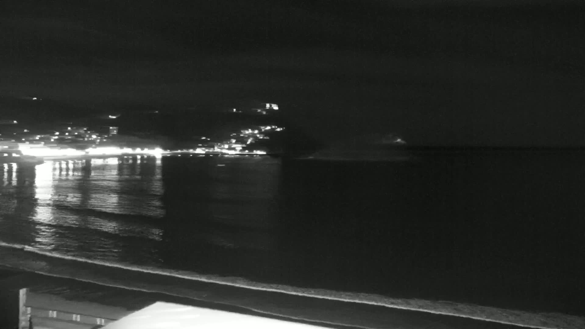 A nighttime view of a calm ocean with a dimly lit town on the shore and dark hills in the background.