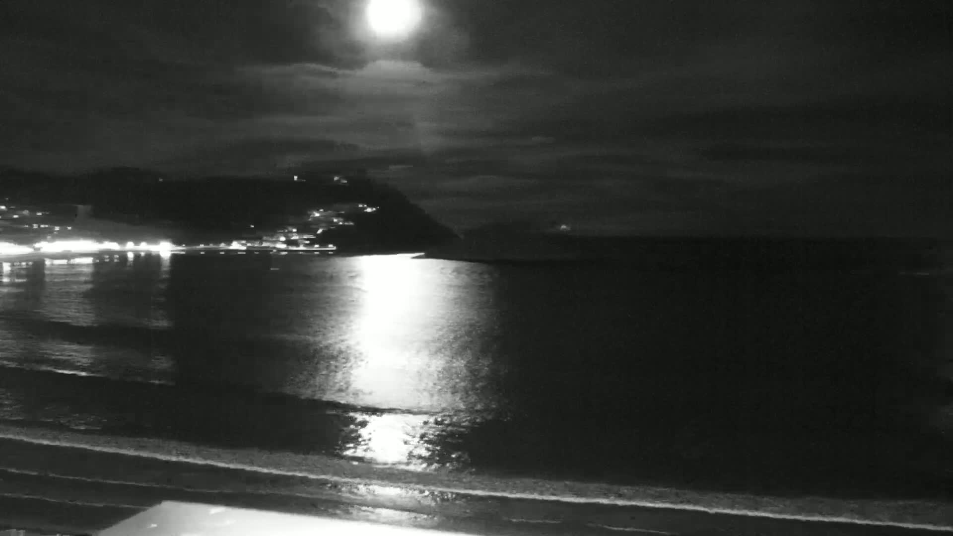 A bright moon illuminates a nighttime coastal scene with calm water reflecting its light, and a dark hillside with lights in the distance.