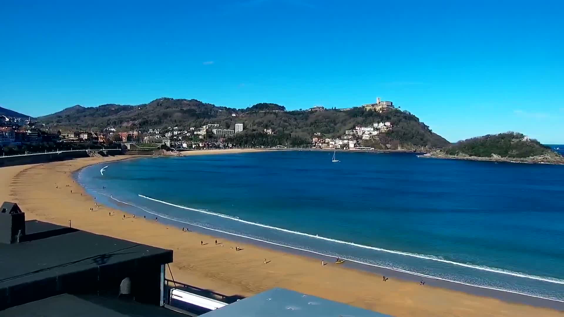A sandy beach curves around a calm, blue bay under a clear, sunny sky, with hills and buildings in the background.