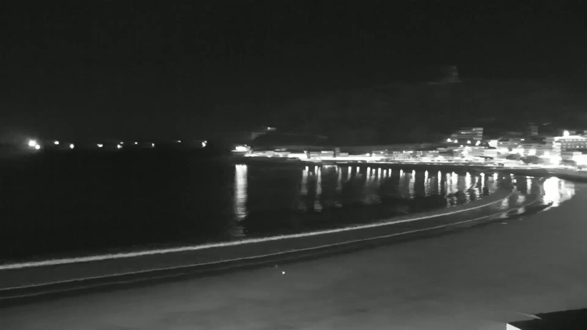 A nighttime view of a dark, sandy beach with gentle waves lapping the shore, illuminated by the lights of buildings across the water.