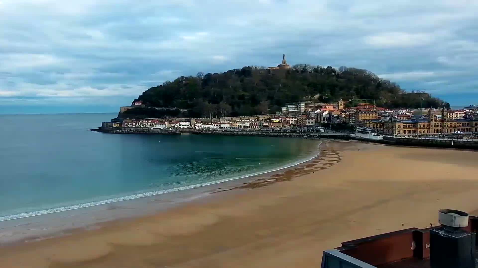 A sandy beach curves along a calm ocean, meeting a town nestled at the base of a hill topped with a statue, all under a cloudy sky.