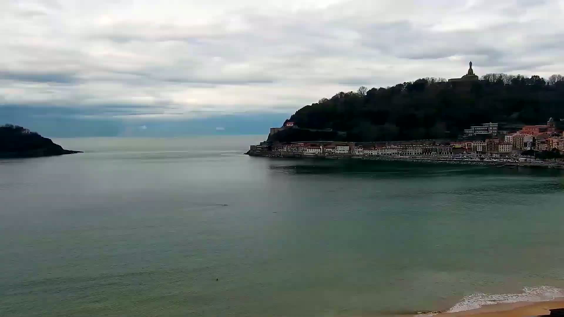A coastal town nestled between two hills sits on a calm bay under a cloudy sky.