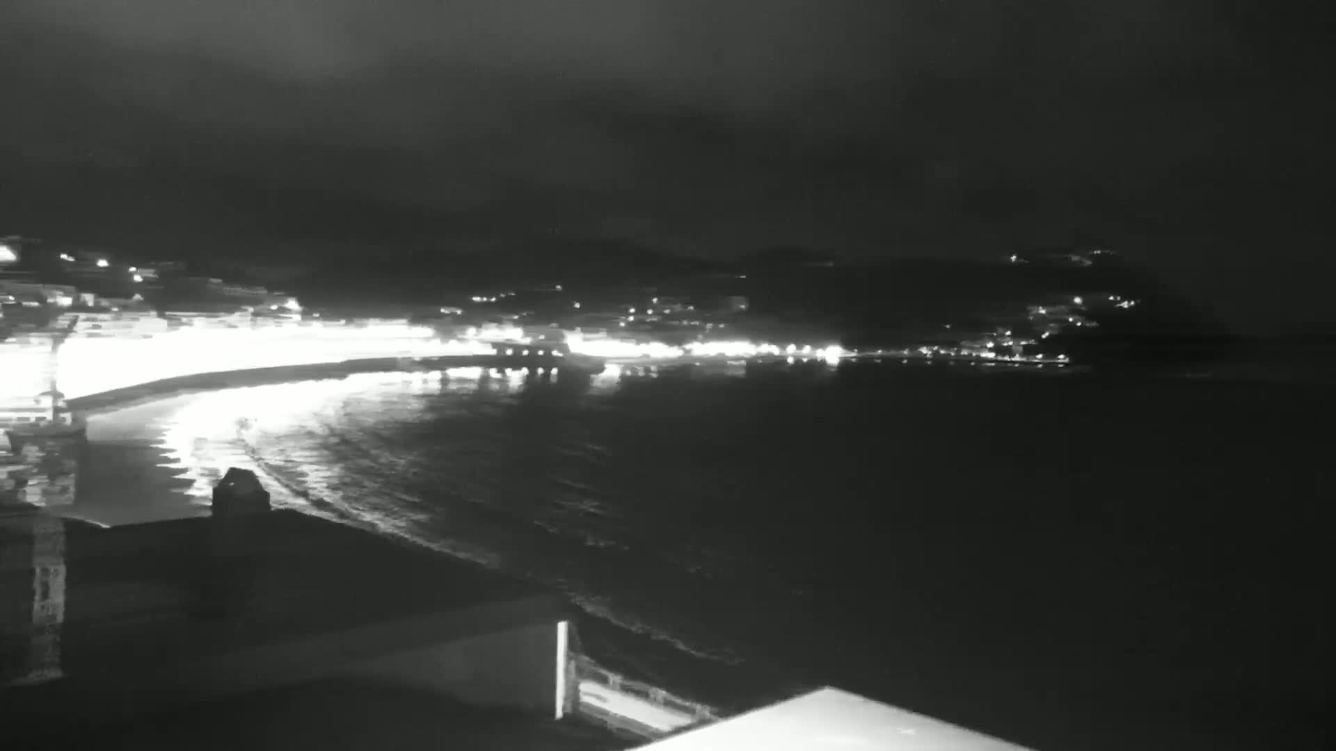 A nighttime view of a calm, dark ocean with a brightly lit coastal town visible in the distance under a cloudy sky.