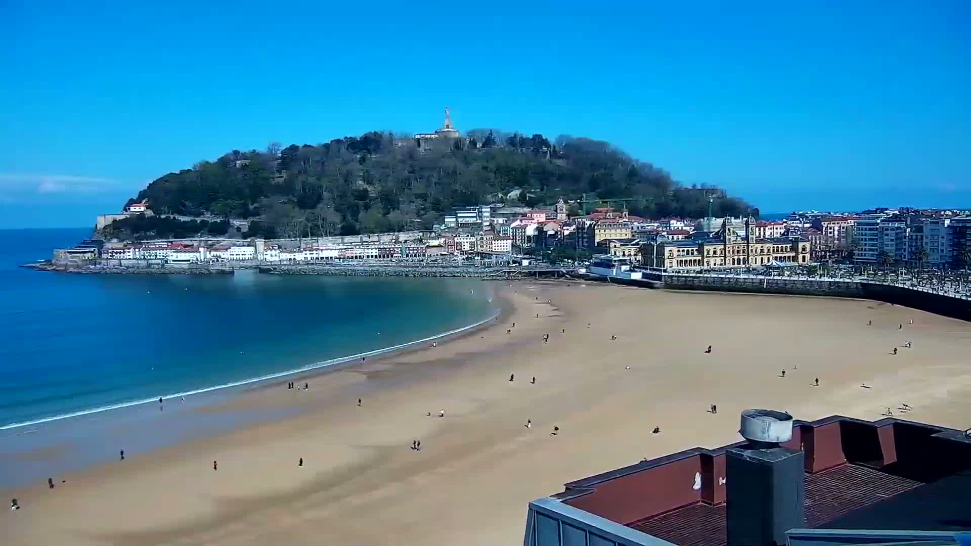 A sandy beach curves around a bay, with a town and a hilltop statue visible under a clear blue sky.