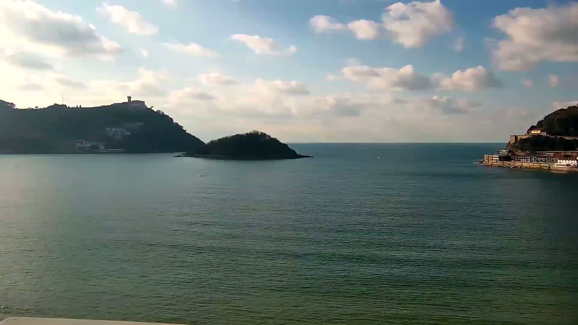 A partly cloudy day reveals a tranquil bay with two hills, one featuring buildings, separated by a small island, and calm dark-green water.