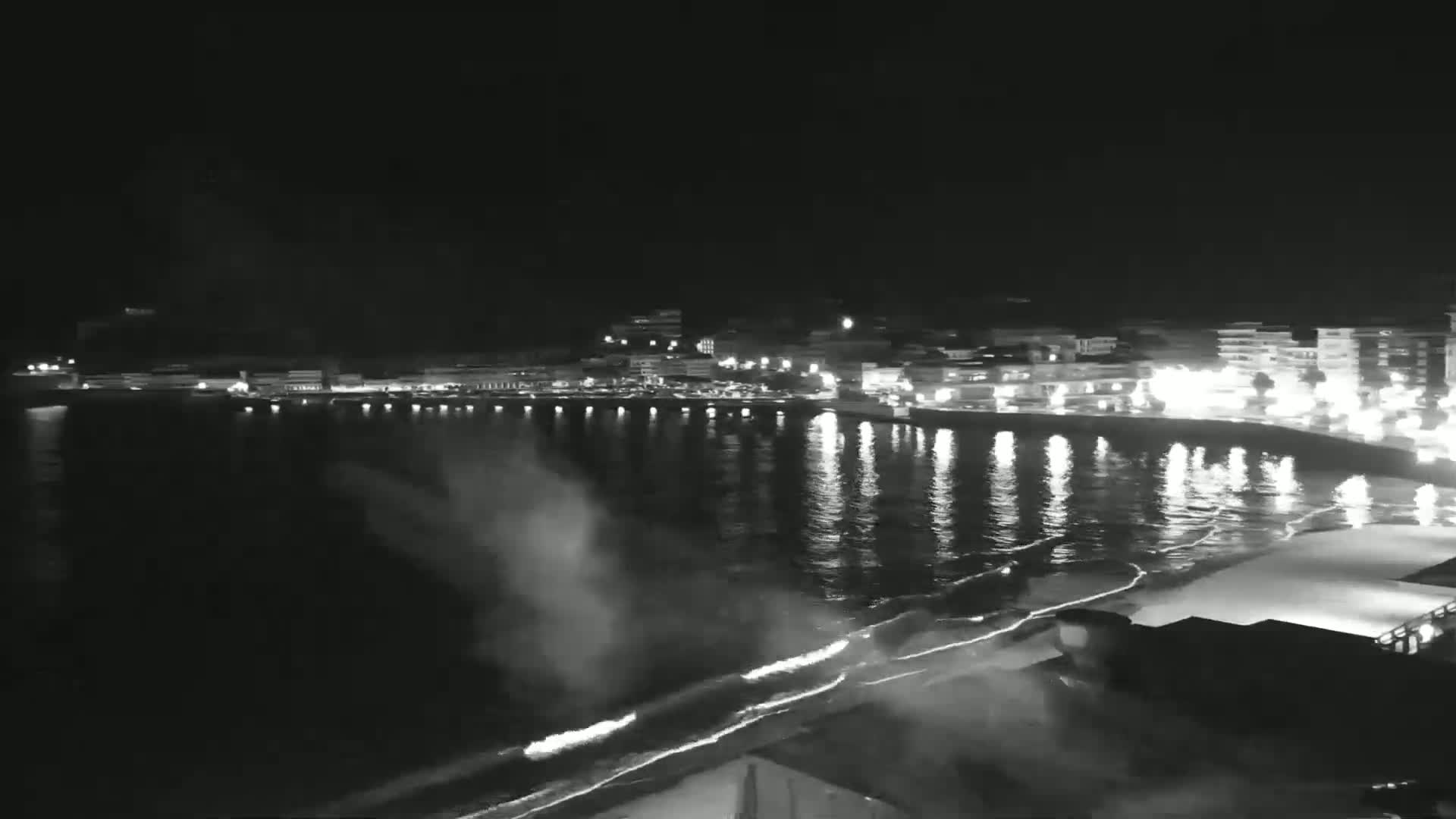 A nighttime, black and white view of a calm ocean reflecting the lights of a town on the shore.