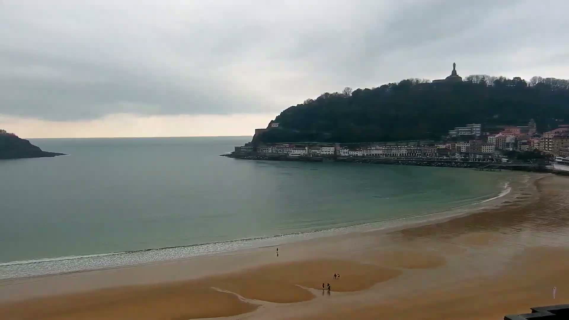 A long, sandy beach curves around a bay under a cloudy sky, with a town nestled at the base of a hill topped by a statue.