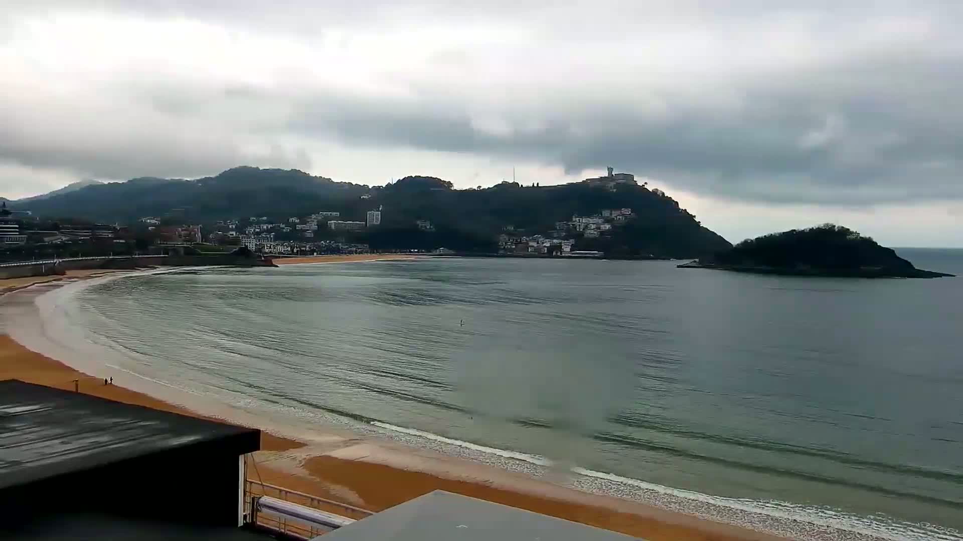 A sandy beach curves into a calm bay under an overcast sky, with a town nestled against hills in the background and a small island off the coast.