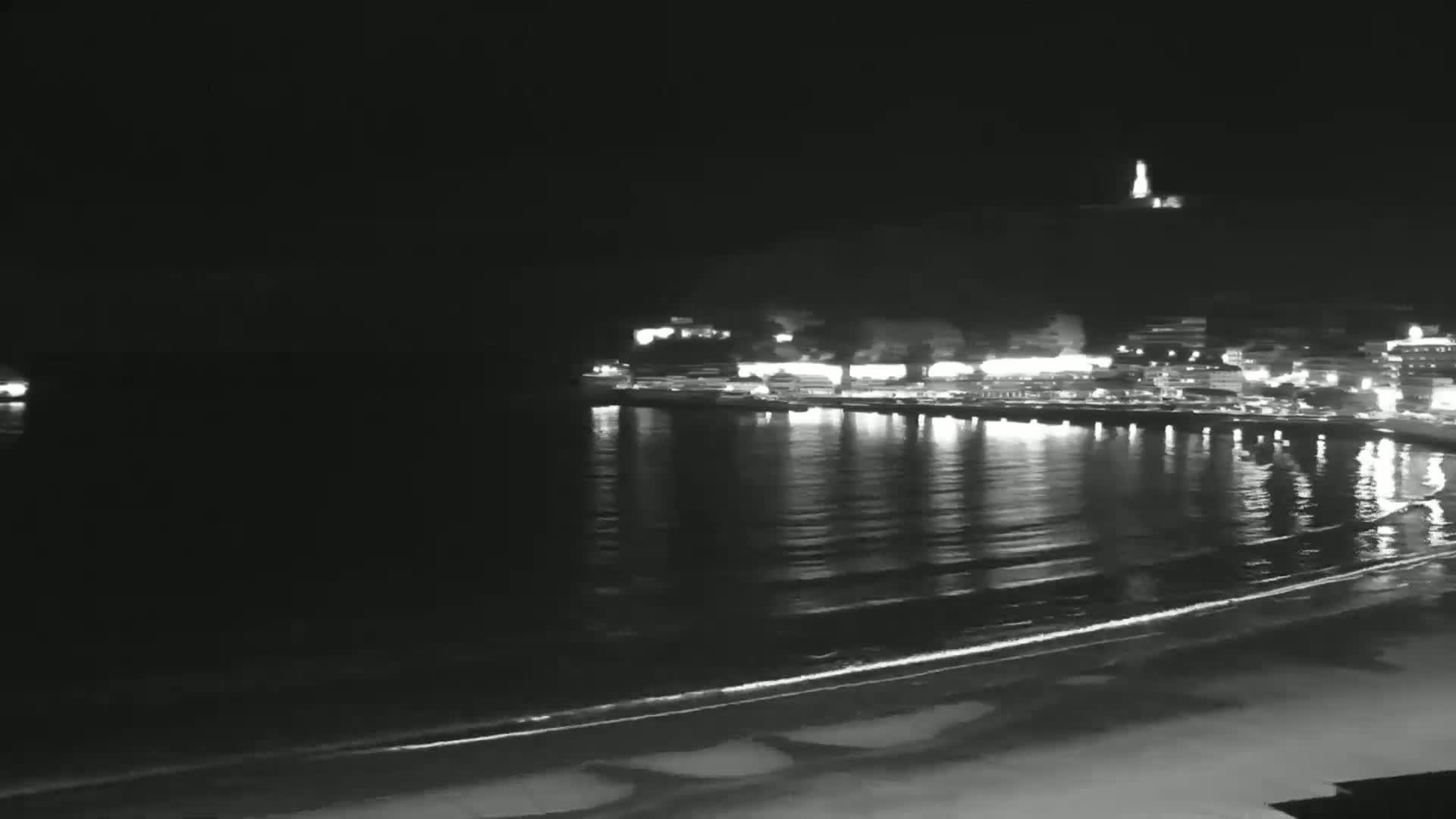 A nighttime view of a calm ocean with a shoreline and a town illuminated on a hill in the background.