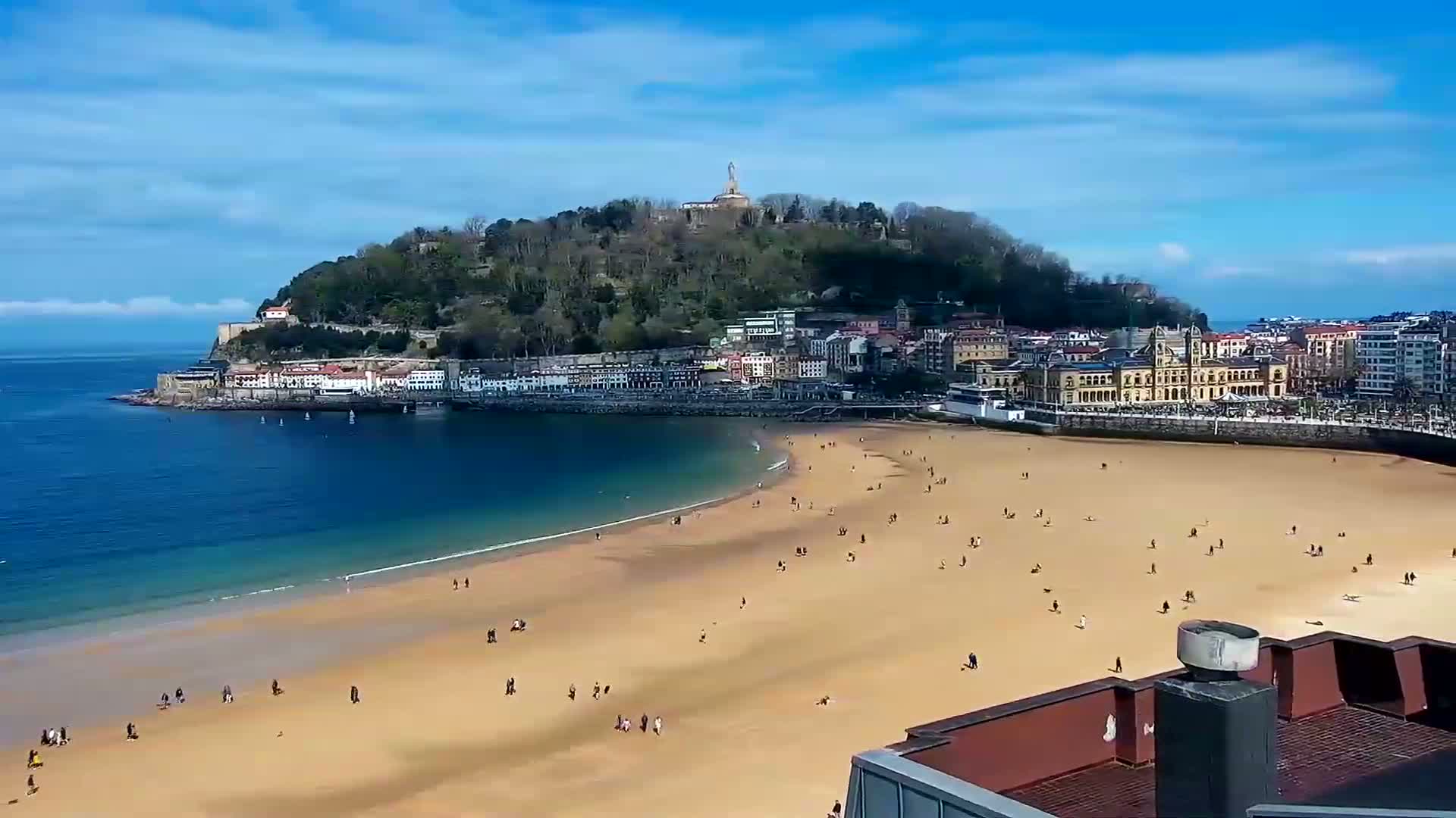 A sandy beach with scattered people stretches along a bay, backed by a town nestled beneath a hill topped with a statue, all under a bright, sunny sky.