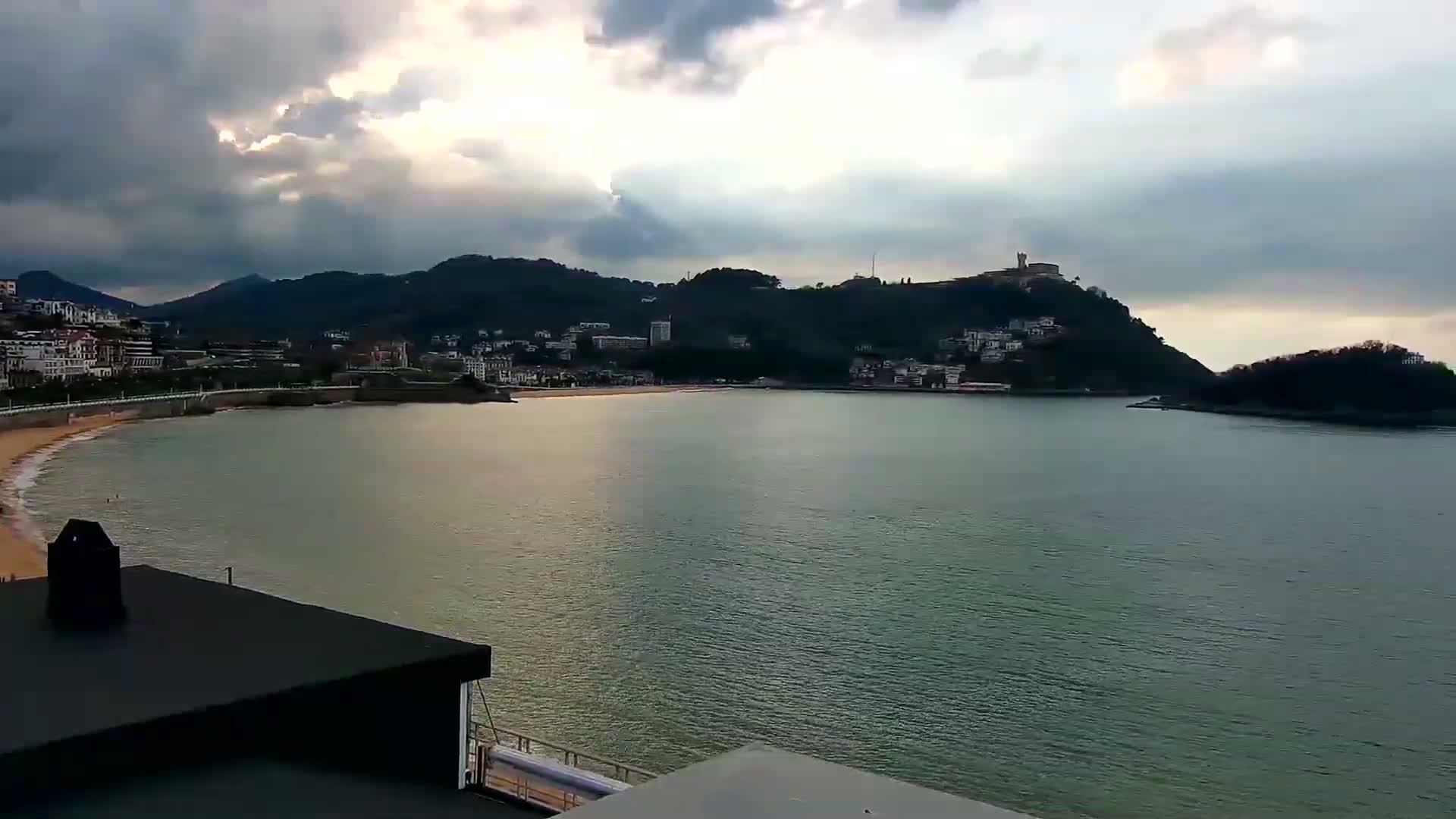 A coastal town nestled between hills and a calm bay under a partly cloudy sky.