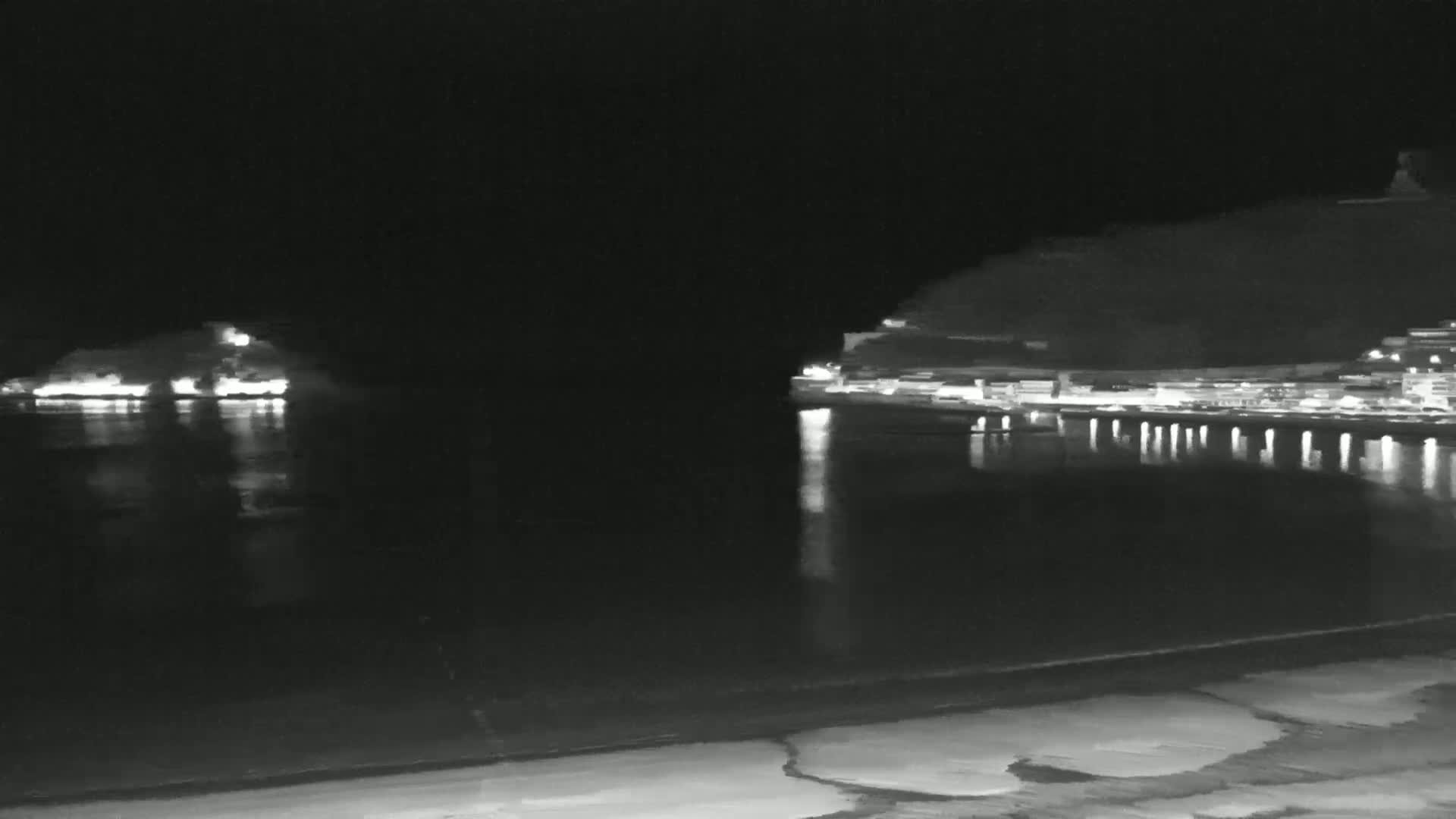 A nighttime view of a dark body of water with lit-up shorelines and hills in the distance, under a clear, dark sky.