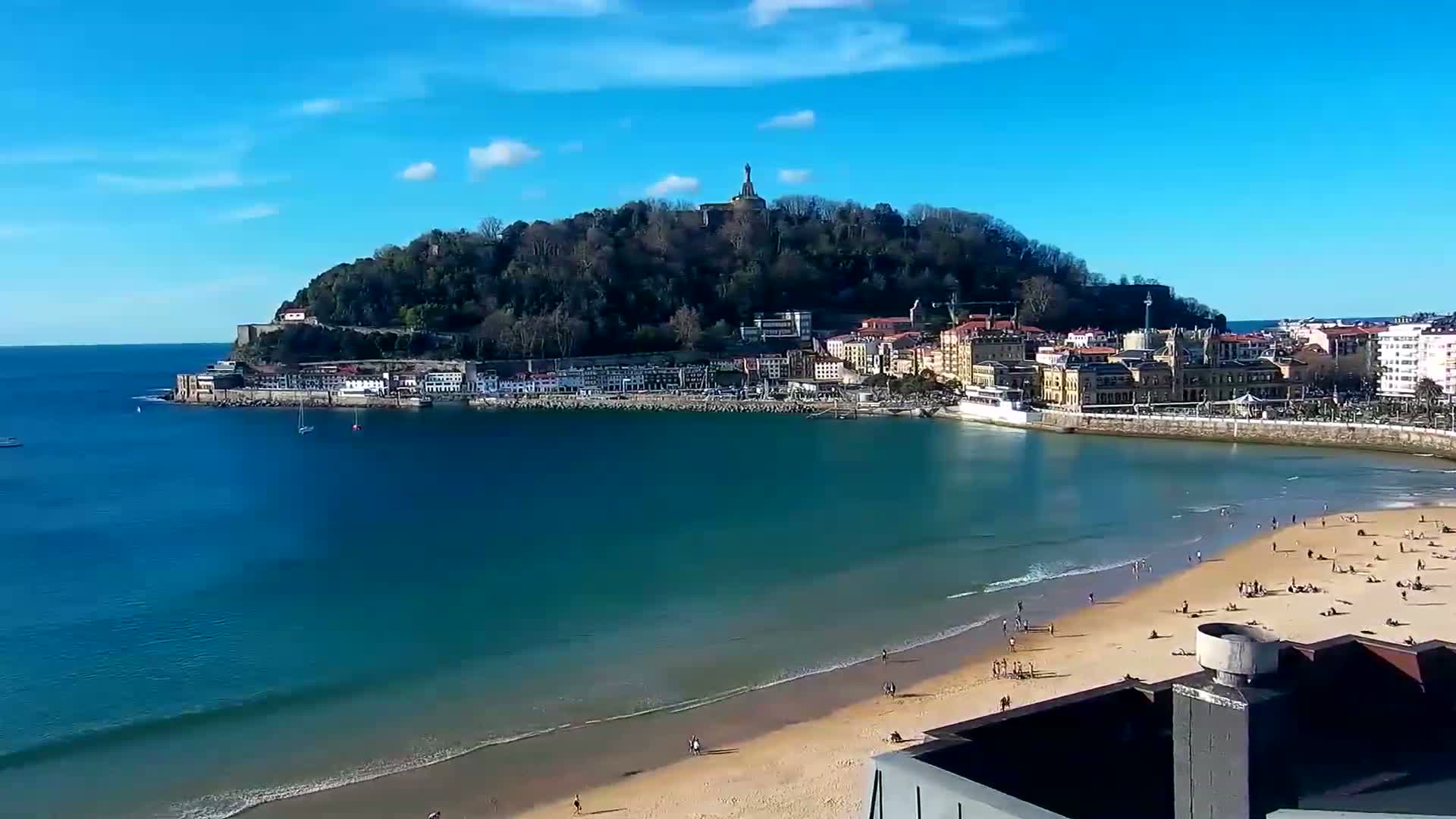 A sandy beach with scattered people is next to calm, teal water that laps a shoreline of buildings nestled at the base of a hill topped with a statue, all under a clear blue sky.
