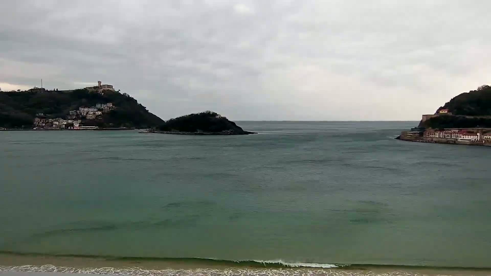 A body of water, under an overcast sky, separates two hillsides, each with buildings along the shoreline, and a small island sits in the middle.