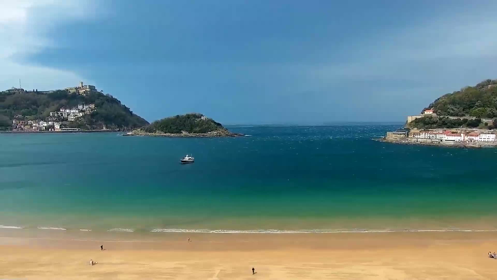 A sandy beach stretches along a calm, teal bay, bordered by hills dotted with buildings under a partly cloudy sky.
