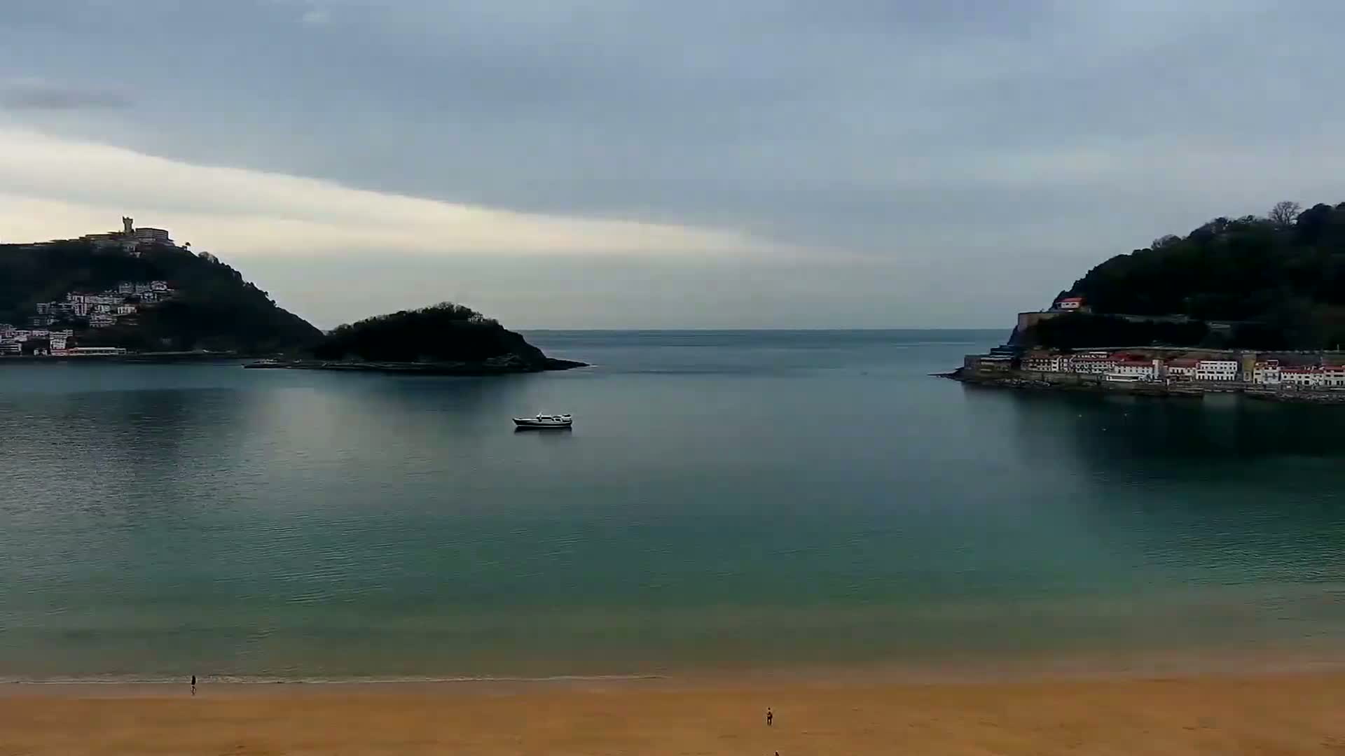 A calm bay with a sandy beach, under an overcast sky, is nestled between two hillsides dotted with buildings.