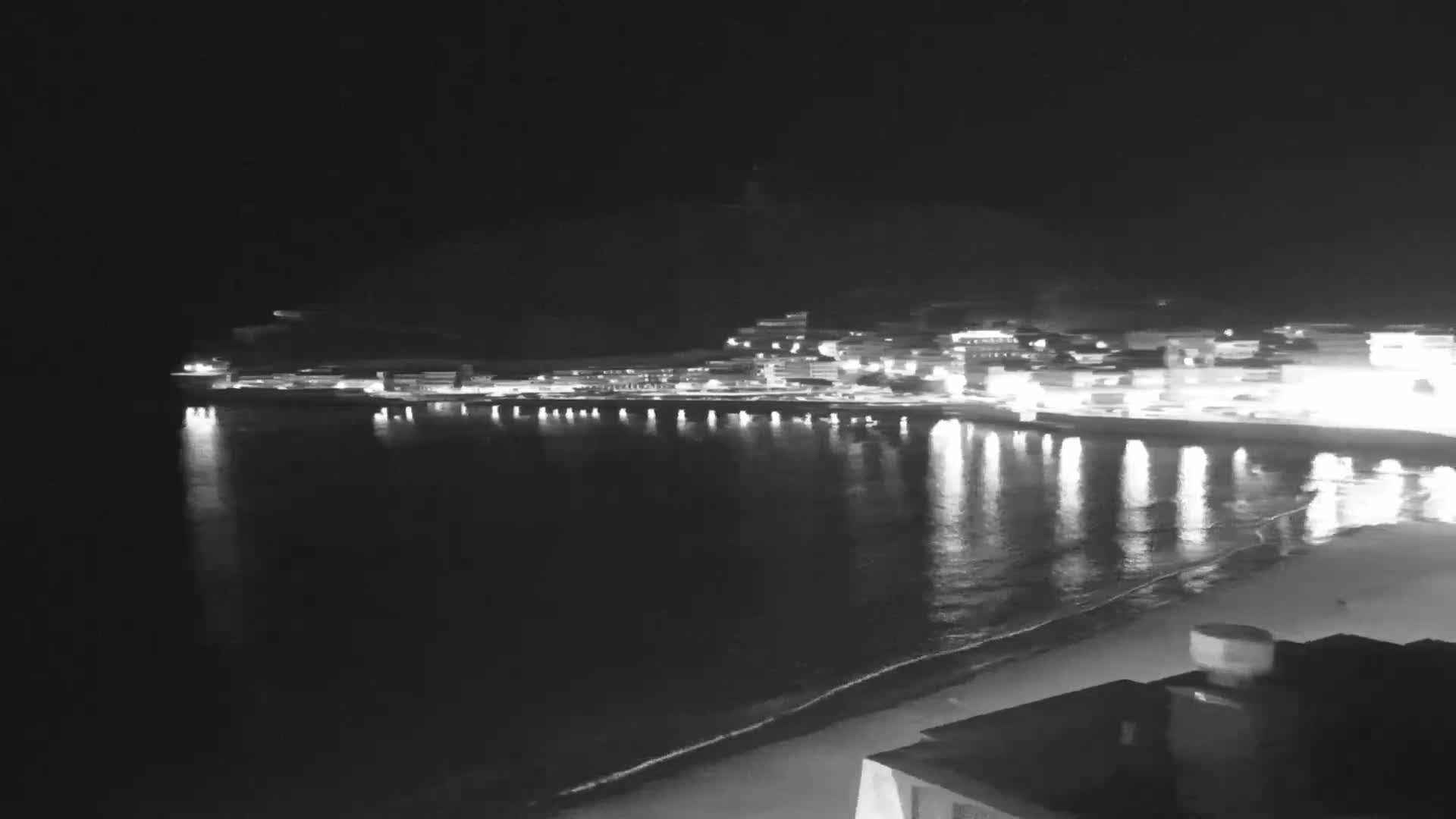 A nighttime view of a town's lights reflected in calm water beside a sandy beach.
