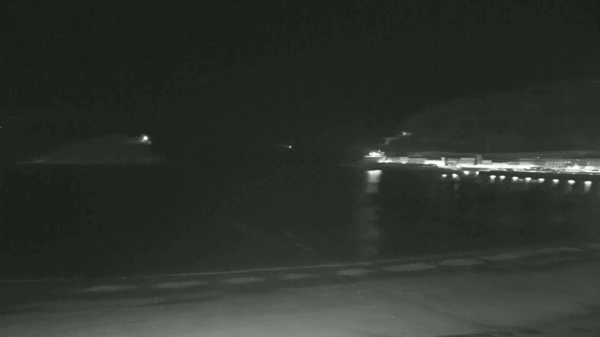 A nighttime view of a dark body of water with dimly lit land and buildings across the water and a dark shoreline in the foreground.
