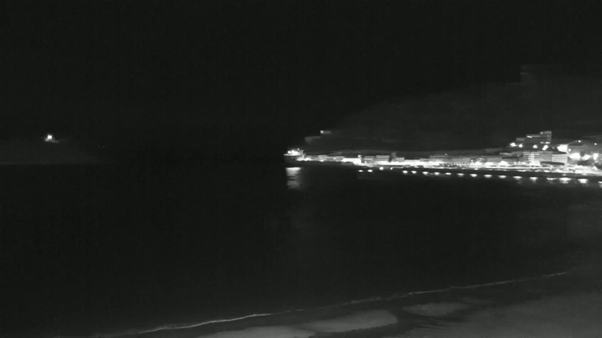 A nighttime view of a dark, mostly calm ocean with a faintly lit shoreline and buildings on a hill in the distance.