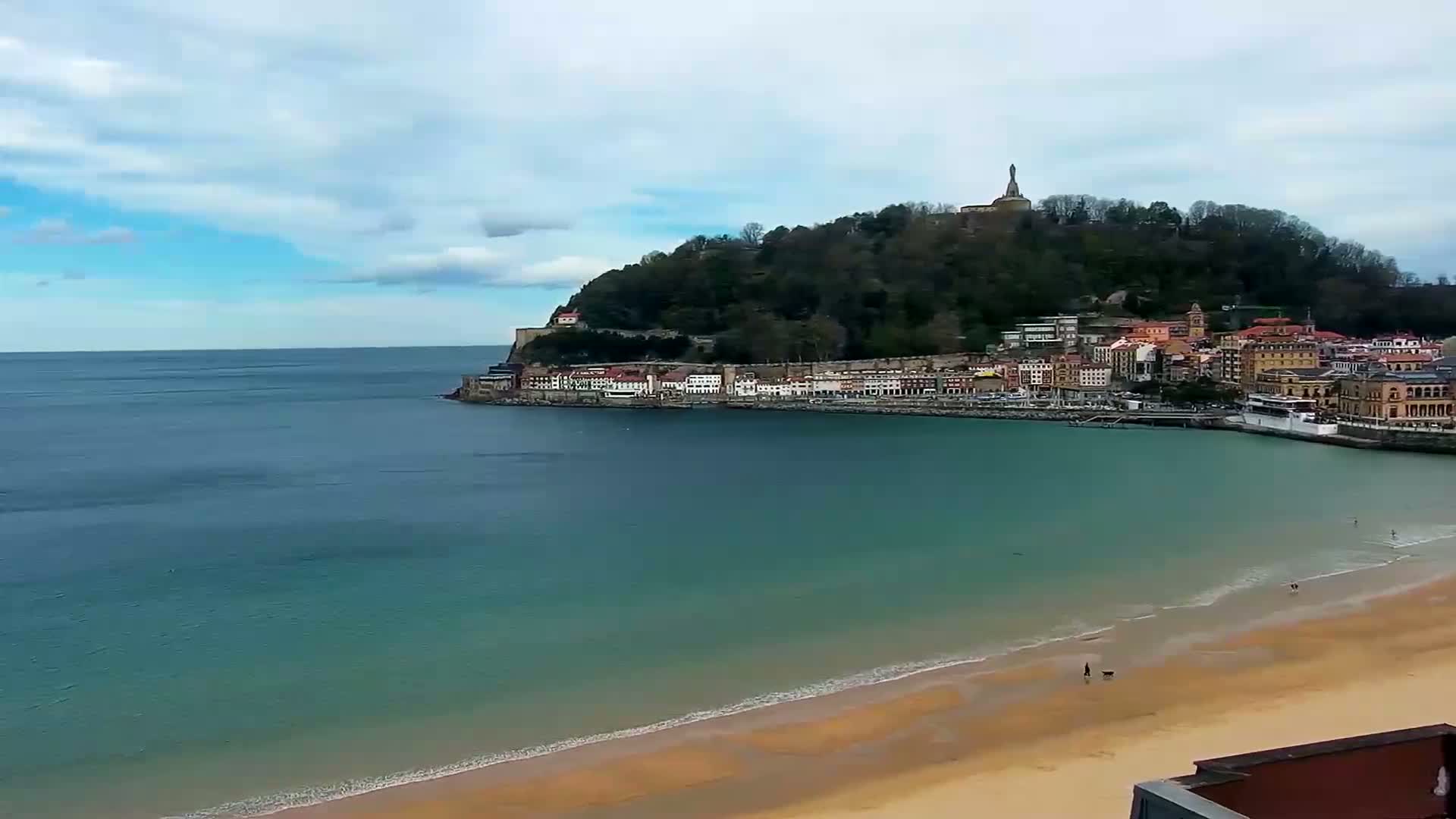 A sandy beach curves into a calm bay, where a town sits nestled at the base of a verdant hill topped with a statue, under a mostly cloudy sky.