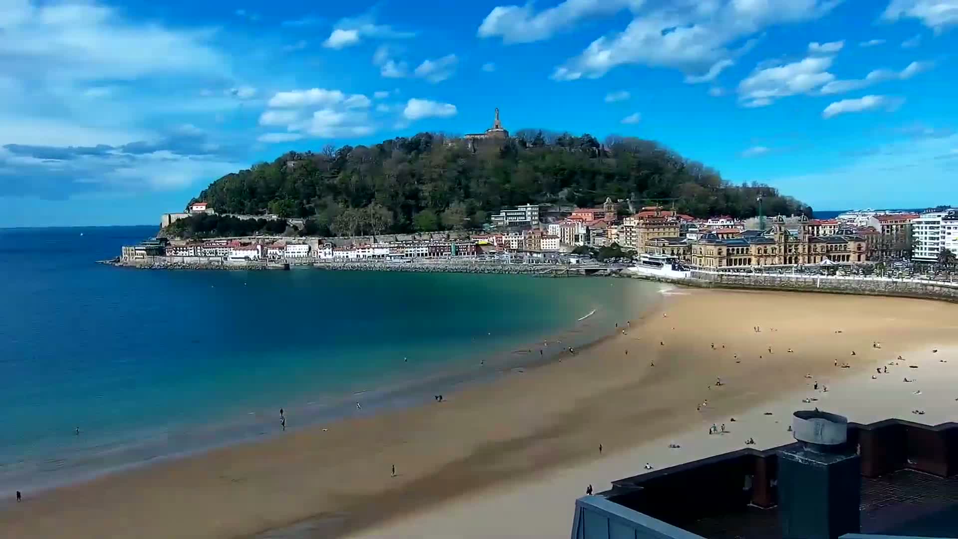 A sandy beach with scattered people stretches along a calm, blue bay, curving around a town nestled beneath a green hill topped with a monument under a mostly sunny sky.