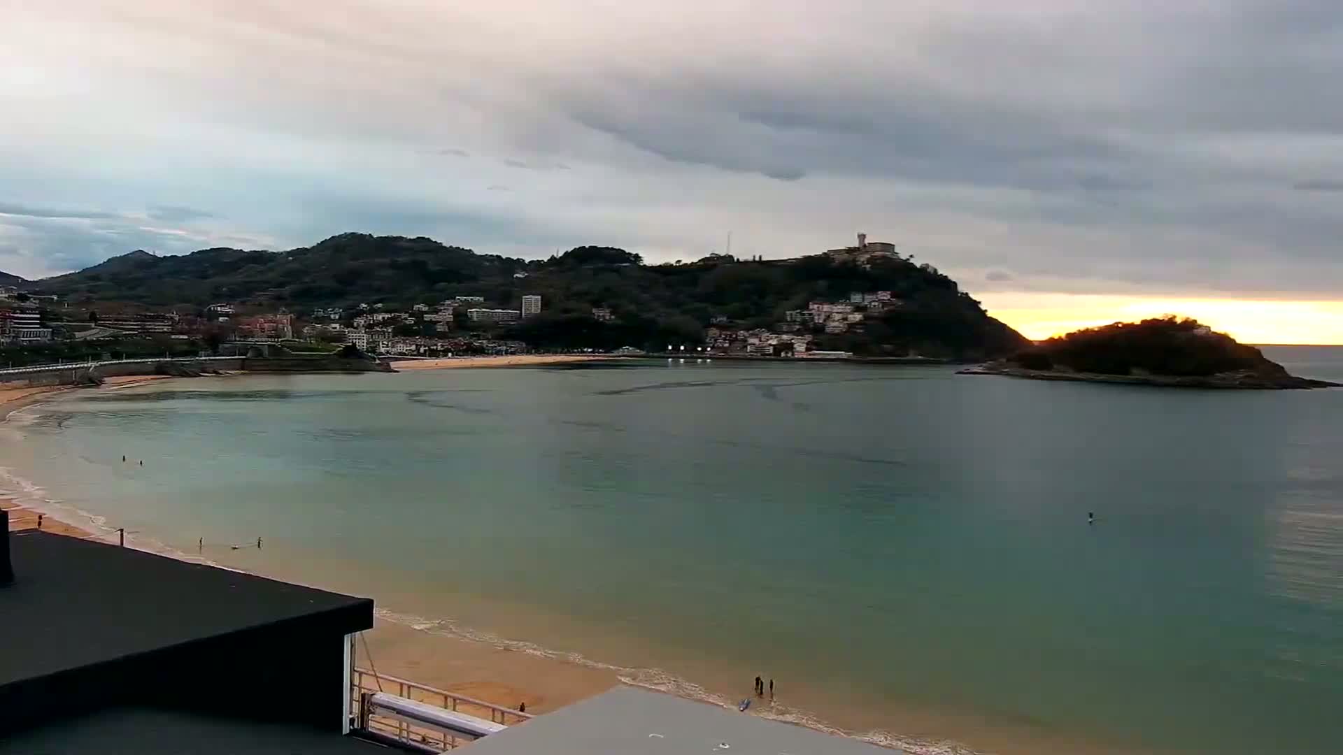 A sandy beach curves around a calm bay under a partly cloudy sky, with hills and buildings rising in the background at sunset.