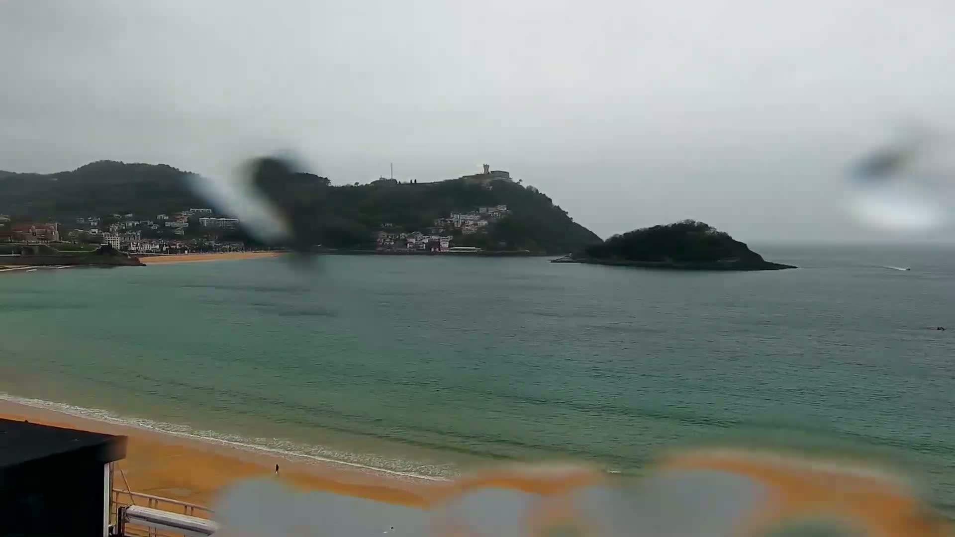 A rain-blurred view shows a sandy beach, calm bay, and a hilltop town nestled against a larger mountain range under an overcast sky.