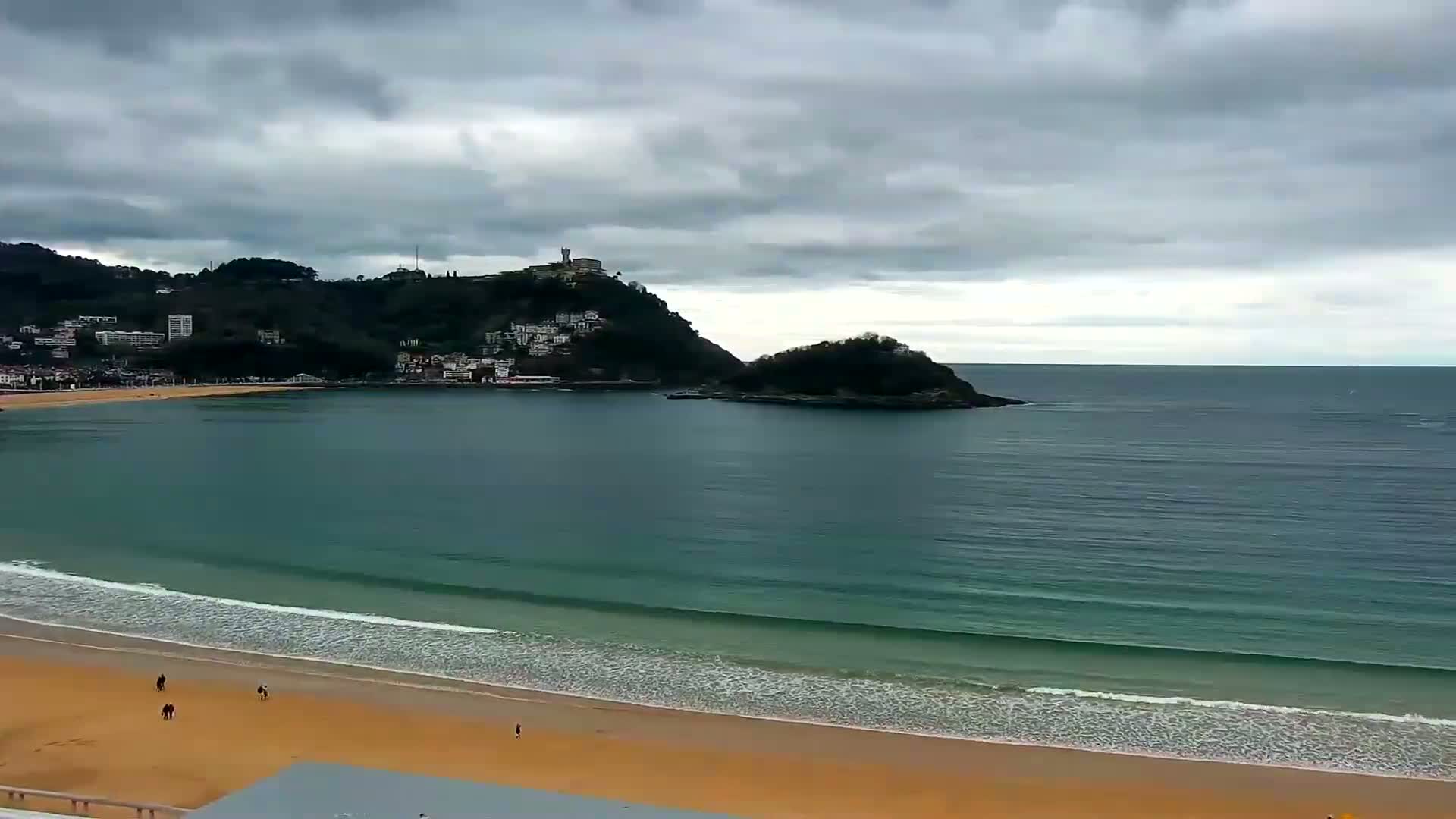 A sandy beach with gentle waves laps the shore under a cloudy sky, with a town nestled at the base of a hill and a small island visible in the bay.