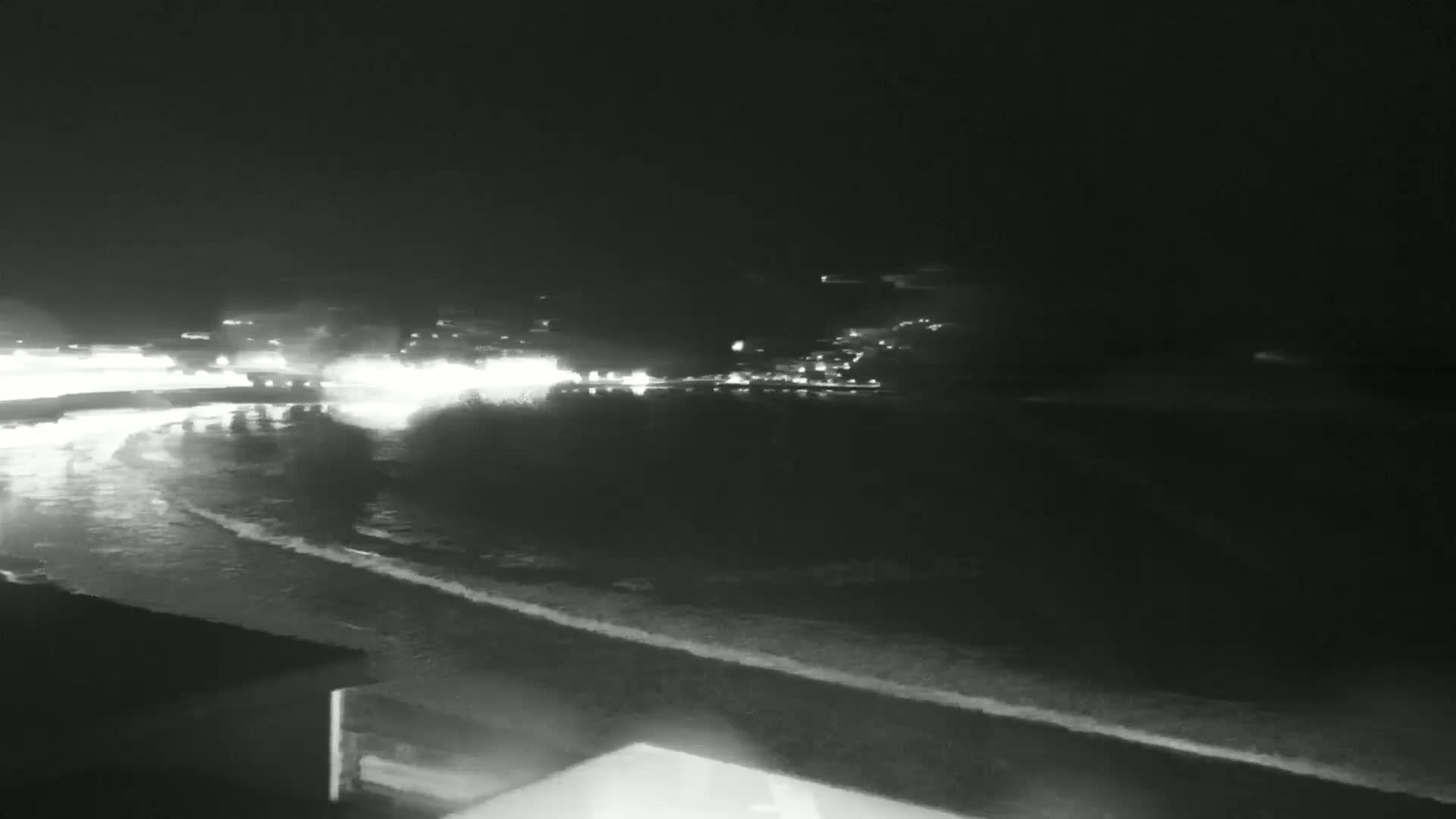 A nighttime, black and white view of a dark ocean shoreline with small waves lapping at the sand, and lights from a town visible in the distance.