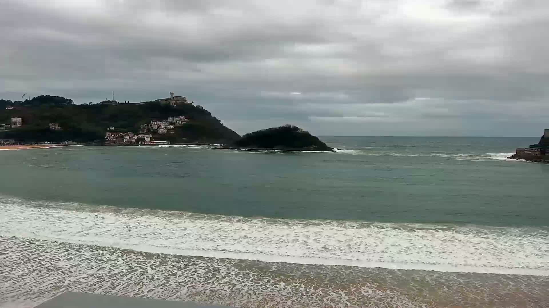 A beach with gentle waves rolls onto the shore under a cloudy sky, with a hill featuring buildings in the distance and a small island in the bay.