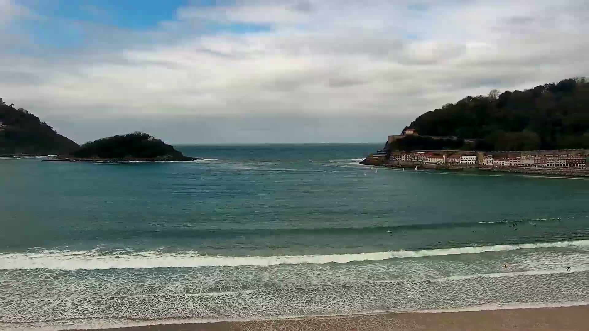 A coastal scene shows a calm bay with a sandy beach, small waves, and a town nestled between two hills under a mostly cloudy sky.