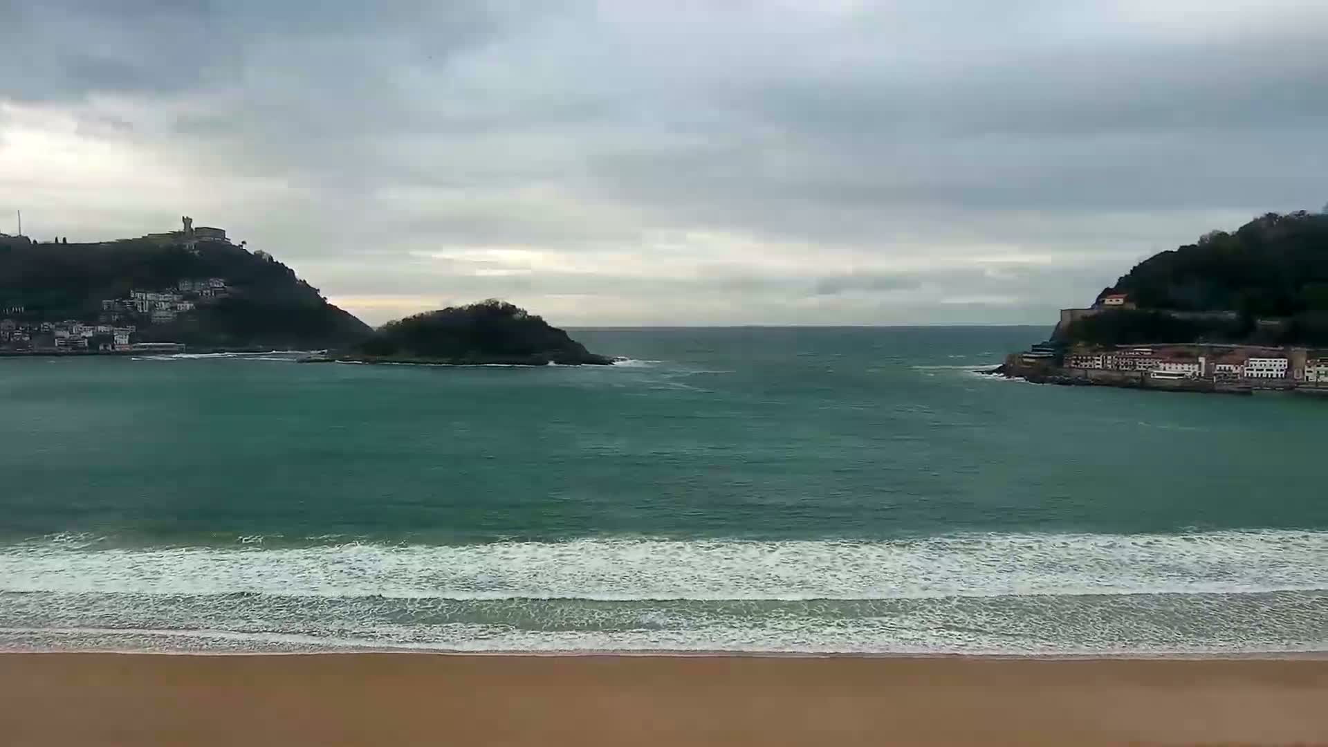 A sandy beach meets a teal ocean with small waves under a cloudy sky, bordered by two hills with buildings on them and a smaller island in between.