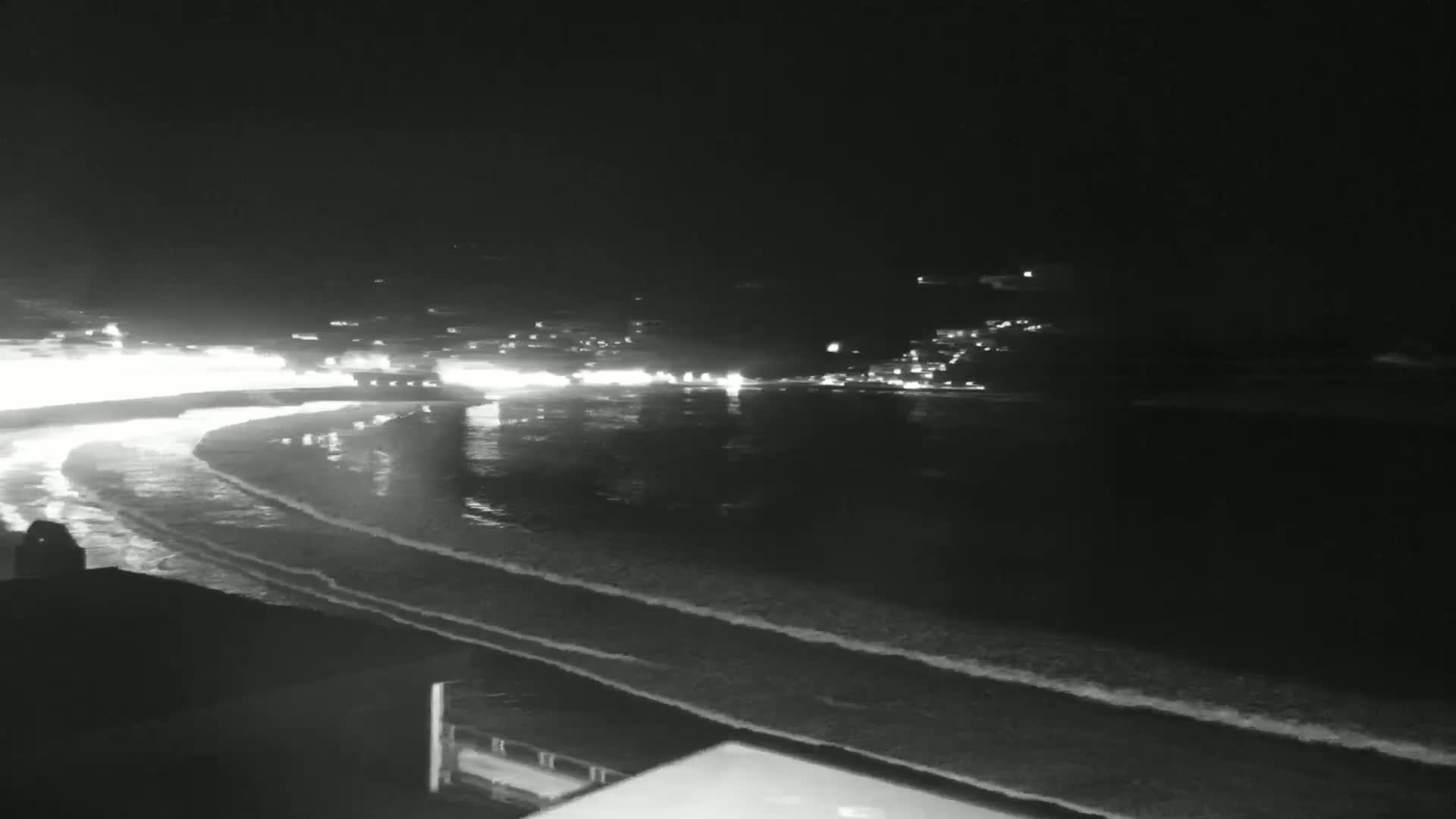 A nighttime view of a dark, wavy ocean shore with a brightly lit town in the distance.
