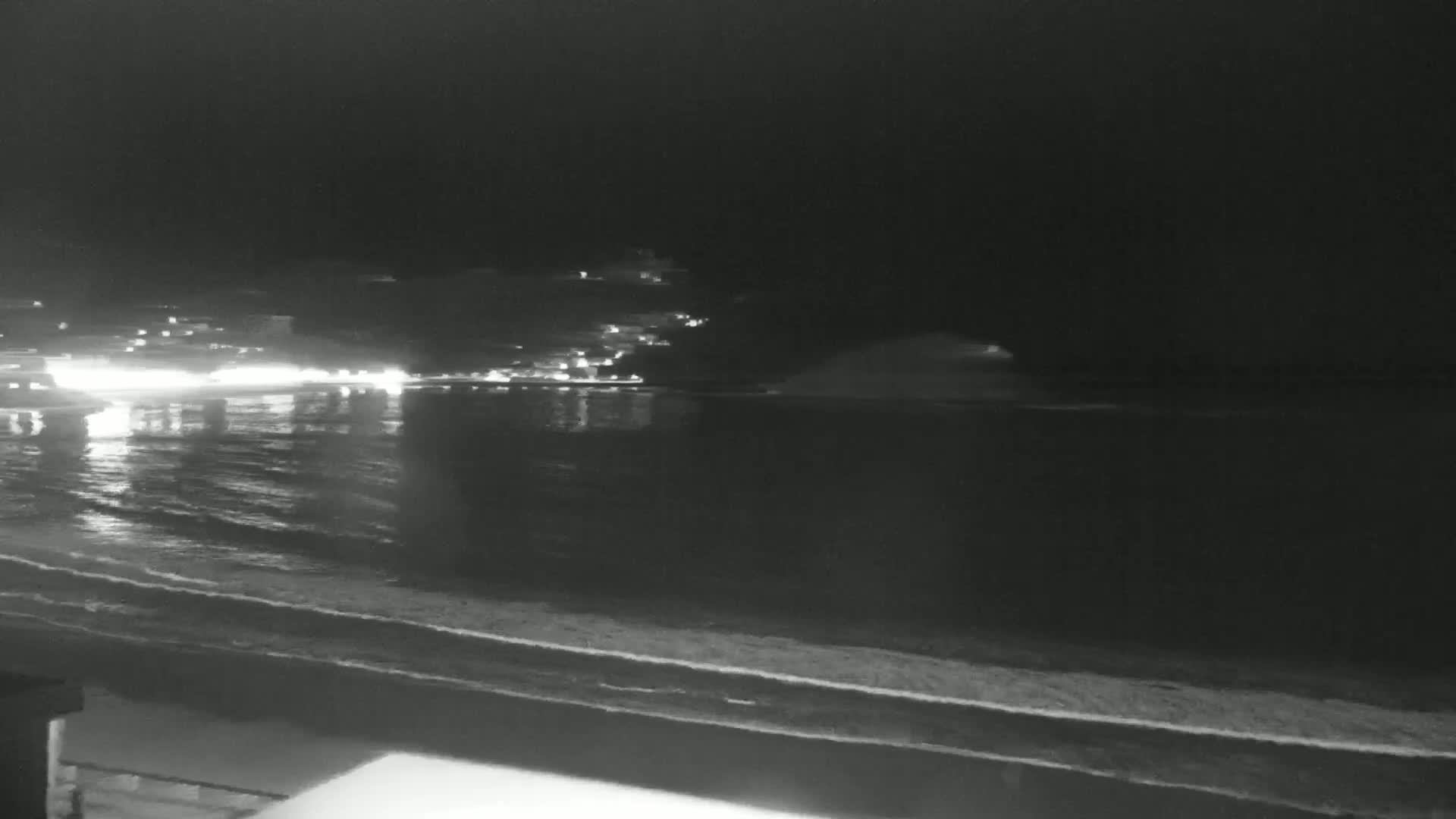 A nighttime view of a dark ocean with gentle waves lapping a shoreline, across from a dimly lit town.