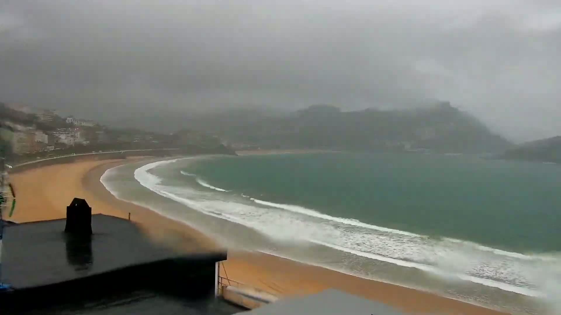 A rain-swept coastal scene shows a curved sandy beach with waves rolling onto the shore, backed by a town nestled against hills shrouded in mist.