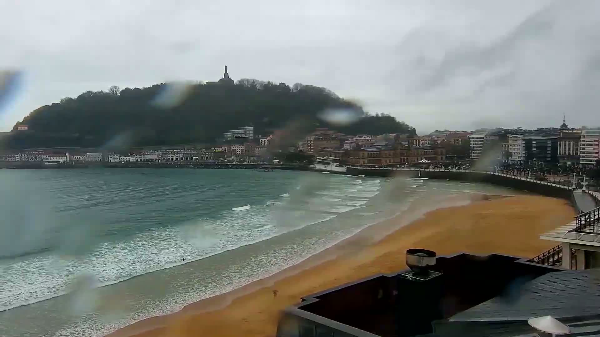 A rain-swept sandy beach curves around a bay, with a town and a hill topped by a monument visible in the distance.