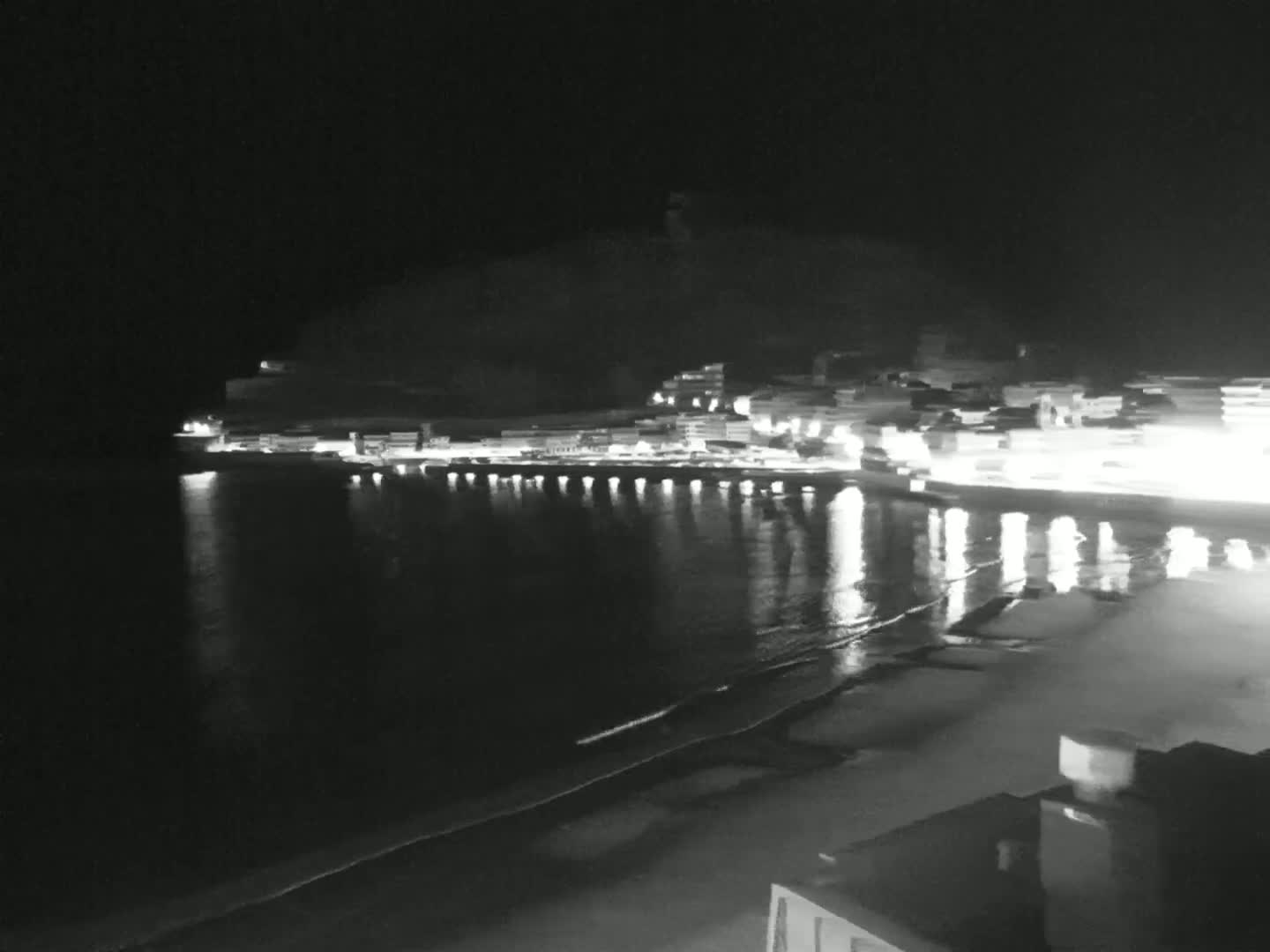 A nighttime view of a calm ocean beach with a town's lights reflected in the water, backed by a hill.