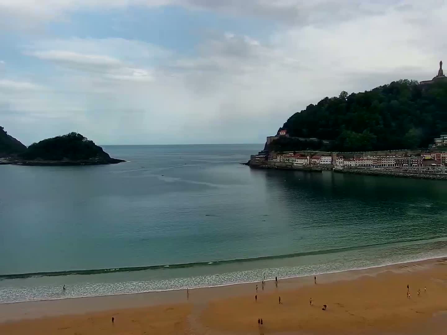 A sandy beach with a few people on it stretches along a calm bay, overlooked by a town nestled at the base of a green hill on an overcast day.