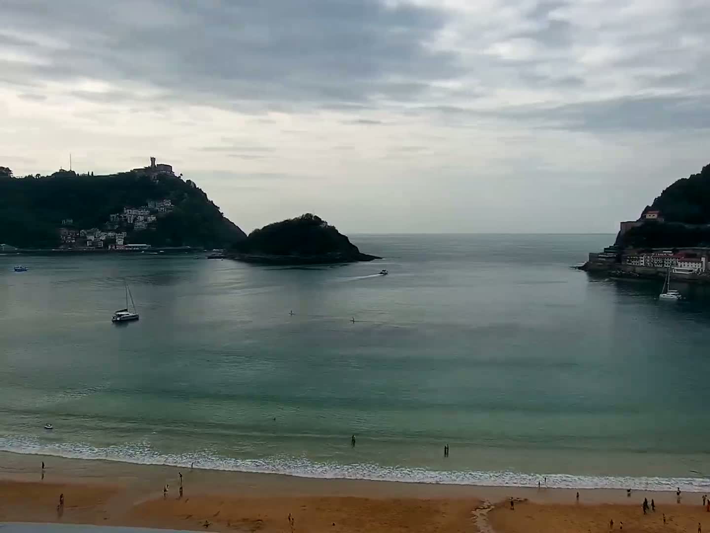 A sandy beach with a calm sea stretches out between two hills, one featuring buildings on its slopes, under an overcast sky.