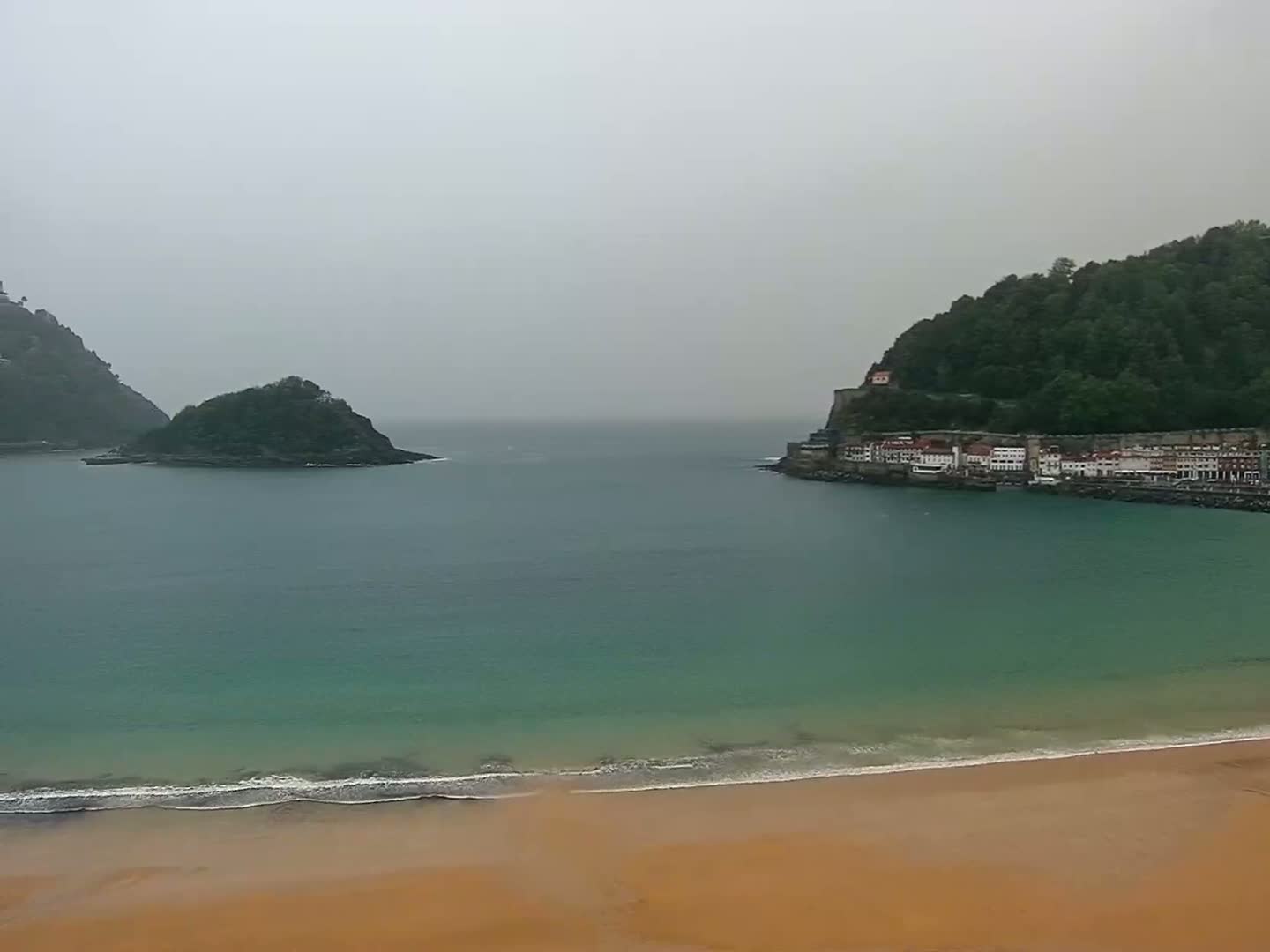A sandy beach borders a calm, teal ocean under an overcast sky, with a small island and a hillside town visible in the distance.