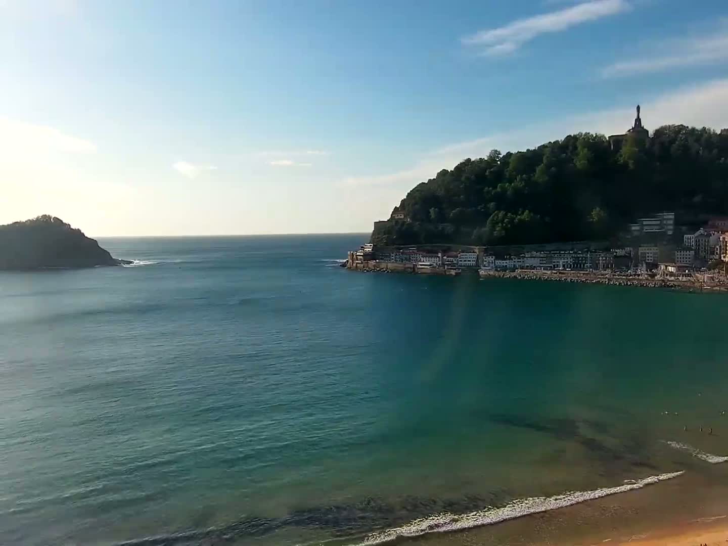 A tranquil coastal scene under a sunny sky shows a town nestled at the base of a verdant hill, with a small island visible across a calm bay.