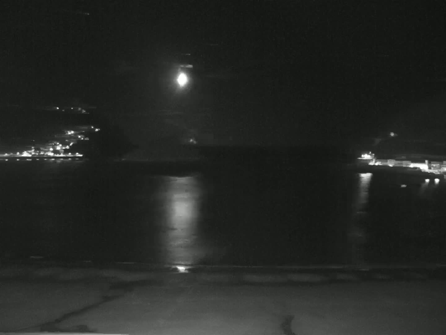 A bright moon illuminates a dark body of water at night, with distant lights visible on either shore.